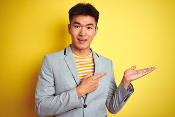 Young asian chinese businessman wearing jacket standing over isolated yellow background amazed and smiling to the camera while presenting with hand and pointing with finger.