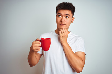 Asian chinese man holding red cup of coffee standing over isolated white background serious face thinking about question, very confused idea