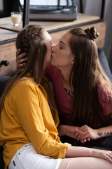 two lesbians kissing and holding hands while sitting on sofa in living room