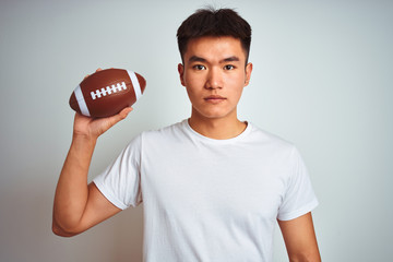 Young asian chinese sportsman holding rugby ball standing over isolated white background with a confident expression on smart face thinking serious