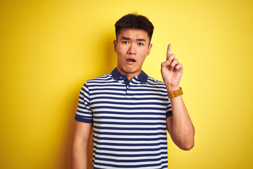 Young asian chinese man wearing striped polo standing over isolated yellow background pointing finger up with successful idea. Exited and happy. Number one.