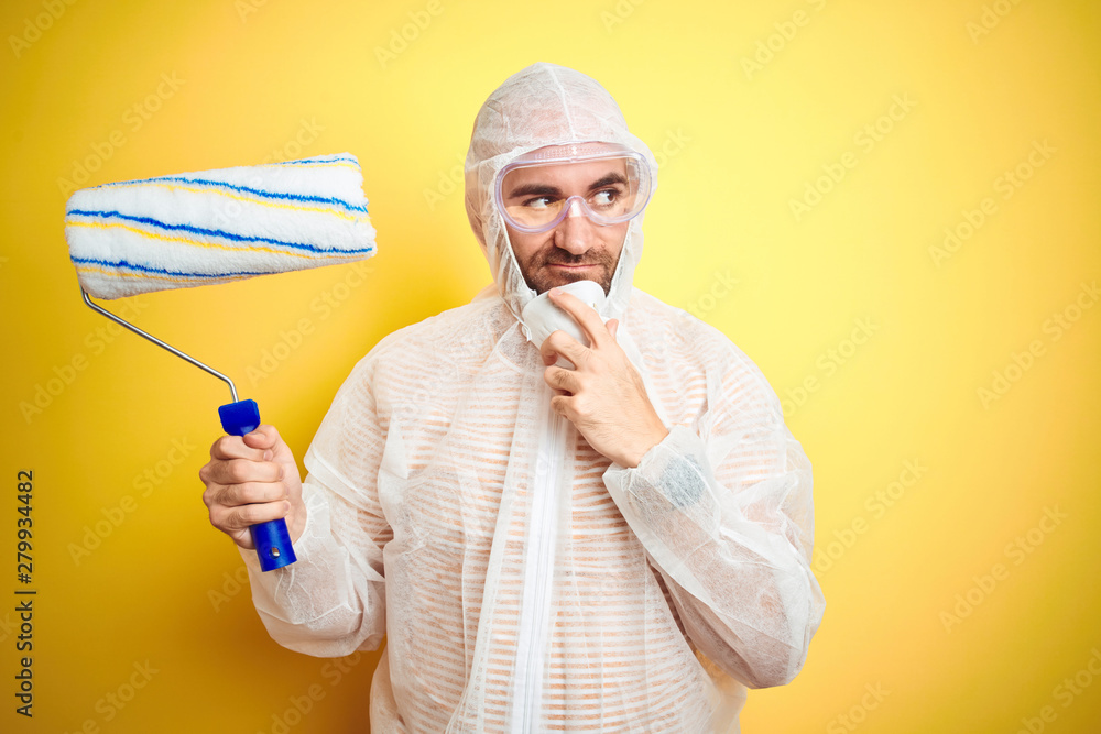 Wall mural Young man wearing painter equipment and holding painting roller over isolated yellow background serious face thinking about question, very confused idea