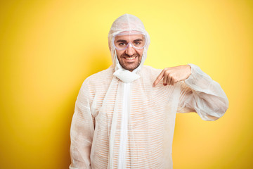 Young man wearing painter equipment and safety glasses over isolated yellow background with surprise face pointing finger to himself