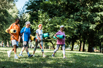 happy retired and multicultural pensioners holding fitness mats and walking in park