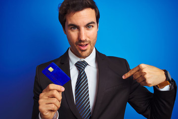 Young handsome businessman holding credit card standing over isolated blue background with surprise face pointing finger to himself