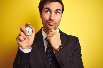 Young handsome businessman holding stopwatch standing over isolated yellow background serious face...