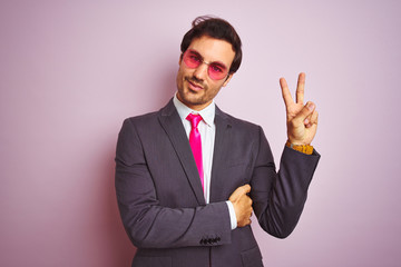 Young handsome businessman wearing suit and sunglasses over isolated pink background smiling with happy face winking at the camera doing victory sign. Number two.