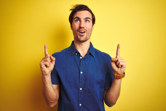 Young handsome man with tattoo wearing casual shirt standing over isolated yellow background amazed and surprised looking up and pointing with fingers and raised arms.