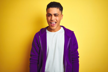 Young brazilian man wearing purple sweatshirt standing over isolated yellow background winking...