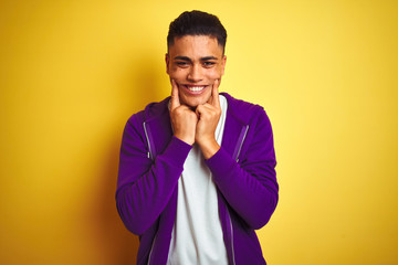 Young brazilian man wearing purple sweatshirt standing over isolated yellow background Smiling with open mouth, fingers pointing and forcing cheerful smile