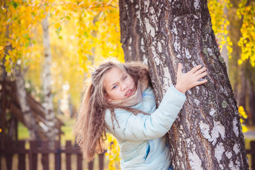 Cute girl with long hair in blue down jacket hugging birch with her hands on an autumn day.