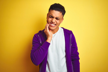 Young brazilian man wearing purple sweatshirt standing over isolated yellow background touching mouth with hand with painful expression because of toothache or dental illness on teeth. 