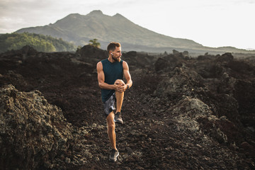 Young hipster runner with beard stretching and warming-up for trail running outdoors. Listening music in air pods. Mountain view on background.