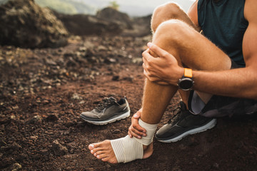 Man bandaging injured ankle. Injury leg while running outdoors. First aid for sprained ligament or tendon. Close-up on dark background.