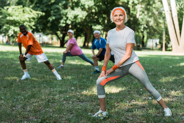 selective focus of cheerful retired woman stretching near multicultural pensioners on grass