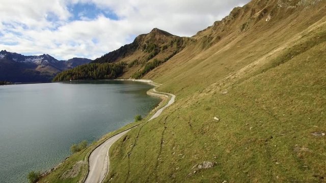 aerial view of mountain road at a lake in Switzerland during autumn with colored pine tree forest, cloudy fall day with snowy mountains, lago ritom