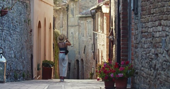 Cheerful attractive girl walking in beautiful narrow street in San Gimignano, Italy. Back view of female photographer traveler taking pictures of italian architecture. Gorgeous cityscape.
