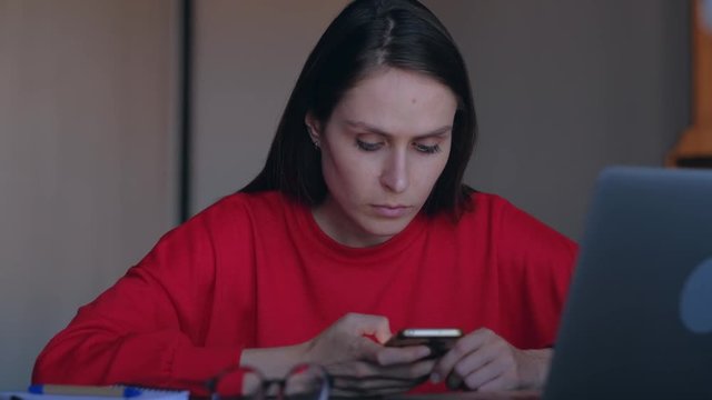 Girl student with red sweatshirt overwrites in smartphone information from the laptop.