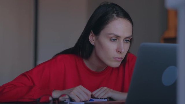 Concept of home education. Cute woman with red sweatshirt overwrites in notepad information from the laptop.