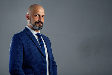Brutal bald bearded businessman in a classic suit on a gray background with copy space