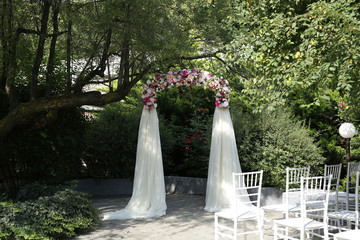 A wedding arch of multicolored flowers and roses with aerial silk fabric for the bride and groom stands under a green tree. Wedding ceremony outdoors. Wedding concept. Botanical style. Rest in harmony