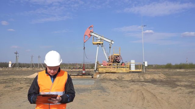 A male inspector-operator in a signal vest and a white helmet records the readings about the pumping unit oil, gas and combustible fuel, industry, copy space, pumpjack, petroleum