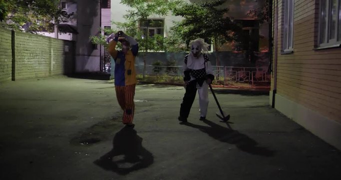 long haired lady runs away from scary clowns in fancy costumes with fake axe and hammer in evening street