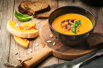 Pumpkin and carrot soup with cream and basil in black plate over wooden background