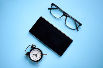 Black glasses, alarm clock and cellphone on blue background composition.