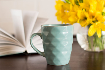 blue coffee mug, yellow flowers in transparent bowl and a book on a wooden table
