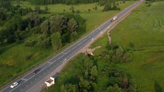 Aerial lateral and pull away shot.Highway with passing cars. Unrecognized truck passing by.The camera flies over the road and green fields and filming highway with traffic signs,old concreet bus stop.