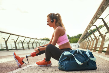 Preparing to run. Side view of young beautiful woman in headphones with leg prosthesis adjusting...