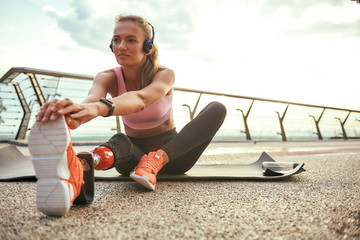 Morning exercises. Young beautiful woman in headphones with leg prosthesis listening music and stretching her leg while sitting on the bridge