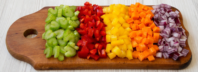Chopped fresh vegetables (carrot, celery, red onion, peppers) arranged on a cutting board on a white wooden background, side view.