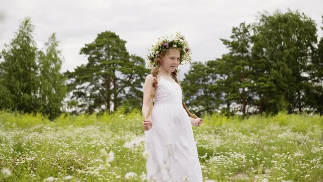Teen girl with daisy wreath and white dress posing for camera in flower meadow. Adorable girl with freckles on her face and long hairs braided in two braids. Summer photo shoot on nature.