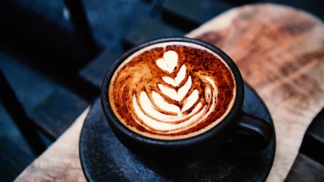 A slight panning shot of cappuccino art. The cup sits on a smooth brown wooden board which is placed on a wooden bench. A lot of dark tones and browns in this which makes the coffee stand out