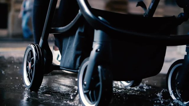 A slow motion shot of a women walking her stroller through a puddle in a city environment, the pram's wheels are splashing the water which is making ripples in the puddle
