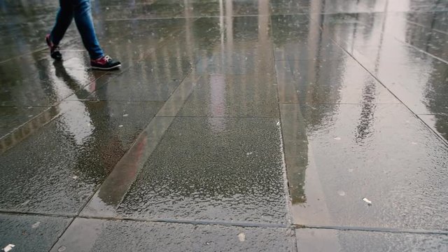 Man In Soaking Wet Blue Jeans And Boat Shoes Walking Through A Rainy Town Centre, The Floor Reflects The Buildings Surrounding This Deserted City. Raindrops Fall Hard And Splash On The Grey Concrete