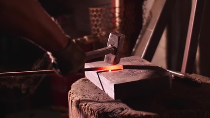 Blacksmith is hitting hot metal bar with a massive hammer on anvil, hands closeup. He is working in smithy workshop in slow motion.