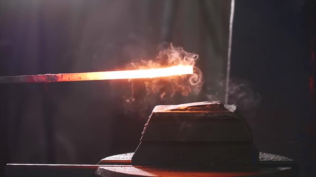Using hammer machine for shaping hot red metal bar in forge workshop, closeup view. Holding beam on a small plate and automatic hammer is hitting on top. Metal forging in metallurgical production.