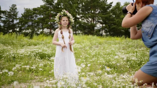 Teen girl is posing and looking at camera on photo shoot. Mom takes pictures of her daughter in white dress with daisy wreath on her head in flower meadow. Nature shot for child girl.