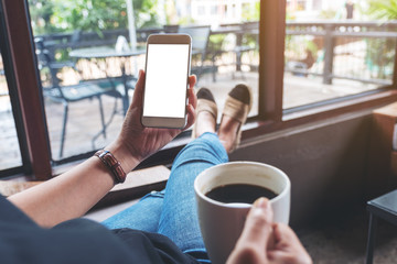 Mockup image of woman holding white mobile phone with blank desktop screen while sitting and drinking coffee in cafe