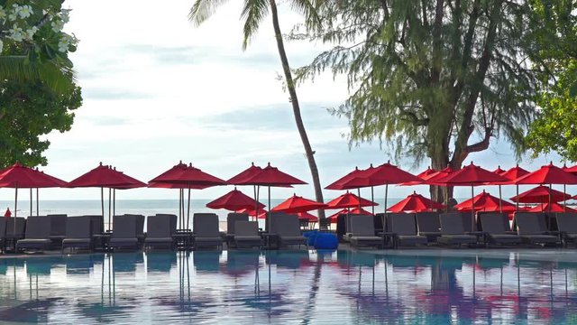 A contrast between clear blue water and a row of red sunshades with gray deckchairs reflects in the infinity pools clam surface