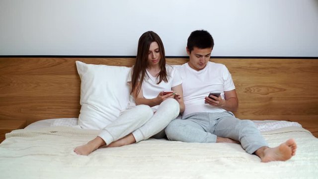 Young couple is sitting in bed and browsing smartphone at home. Happy couple is watching smartphone online content sitting on couch in bright living room at home