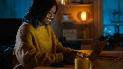 Portrait of Beautiful Black Girl Uses Computer while Sitting at Her Desk at Home, She's Wearing Warm Sweater. In the Evening Creative Woman Works on a Computer In Her Cozy Living Room. 