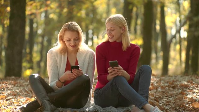 Girls with phone. Young blogger, blog concept. Two girls using smart phone. Online learning, e-learning concept. Beautiful girls in sweater in the autumn nature. Warm Autumn sunny weather.