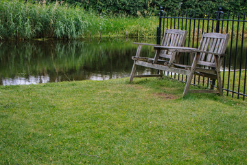 Two wooden chairs in the backyard near pond. Old outdoor furniture in summer garden. Empty quiet place for relax. Dutch patio with retro wooden bench. Lake with lawn and trees. Summer landscape.