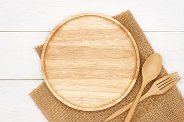 Empty round wooden plate with spoon, fork and rustic brown burlap cloth on white wooden table. Top...