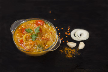 Red lentil soup in a glass plate with spices, herbs, onion and garlic on a black background.