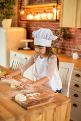 Happy girl 7 years old in chef clothes is standing at the table. A girl in chef clothes sifts flour through a sieve. Homemade food concept.